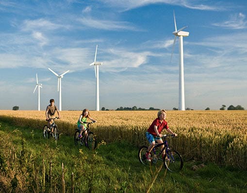 3 children on bicycles in front of wind turbines in a wheat field.