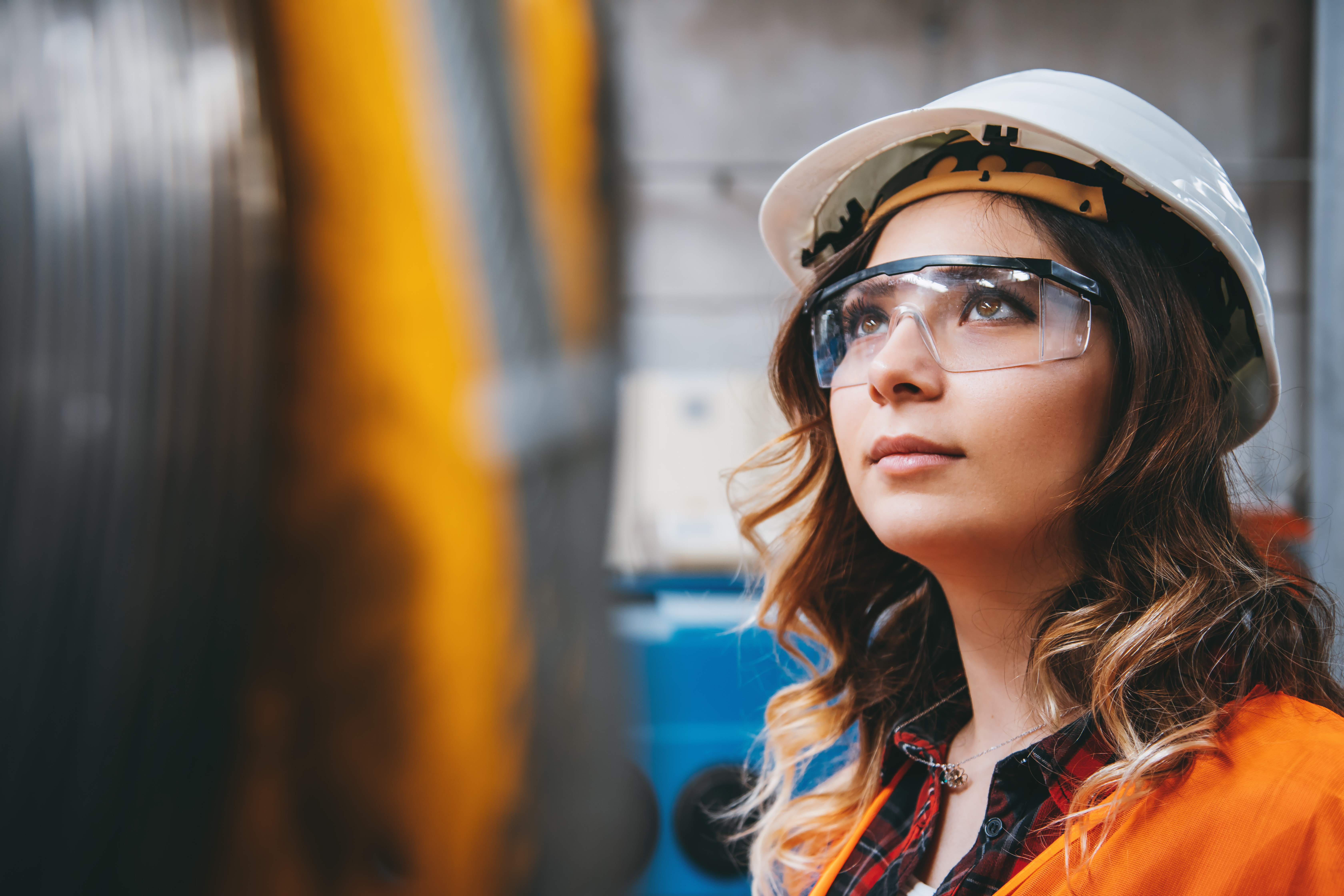 Portrait of young businesswoman with white helmet looking up and seen from the industrial steel cable reel for crane in factory warehouse. She is testing and working with winding wire and cable drum accessories metal wire spool reel wear resistance.